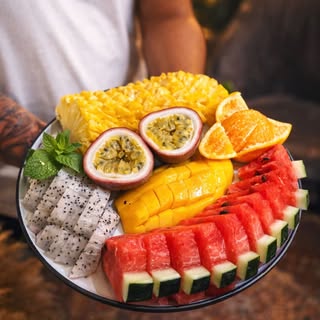 A fresh tropical fruit platter — mango, watermelon, dragon fruit, passion fruit, pineapple — held in two hands.