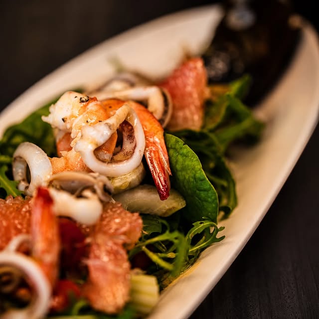 A close-up of a seafood salad: prawn, calamari, rocket, and citrus on a white plate.